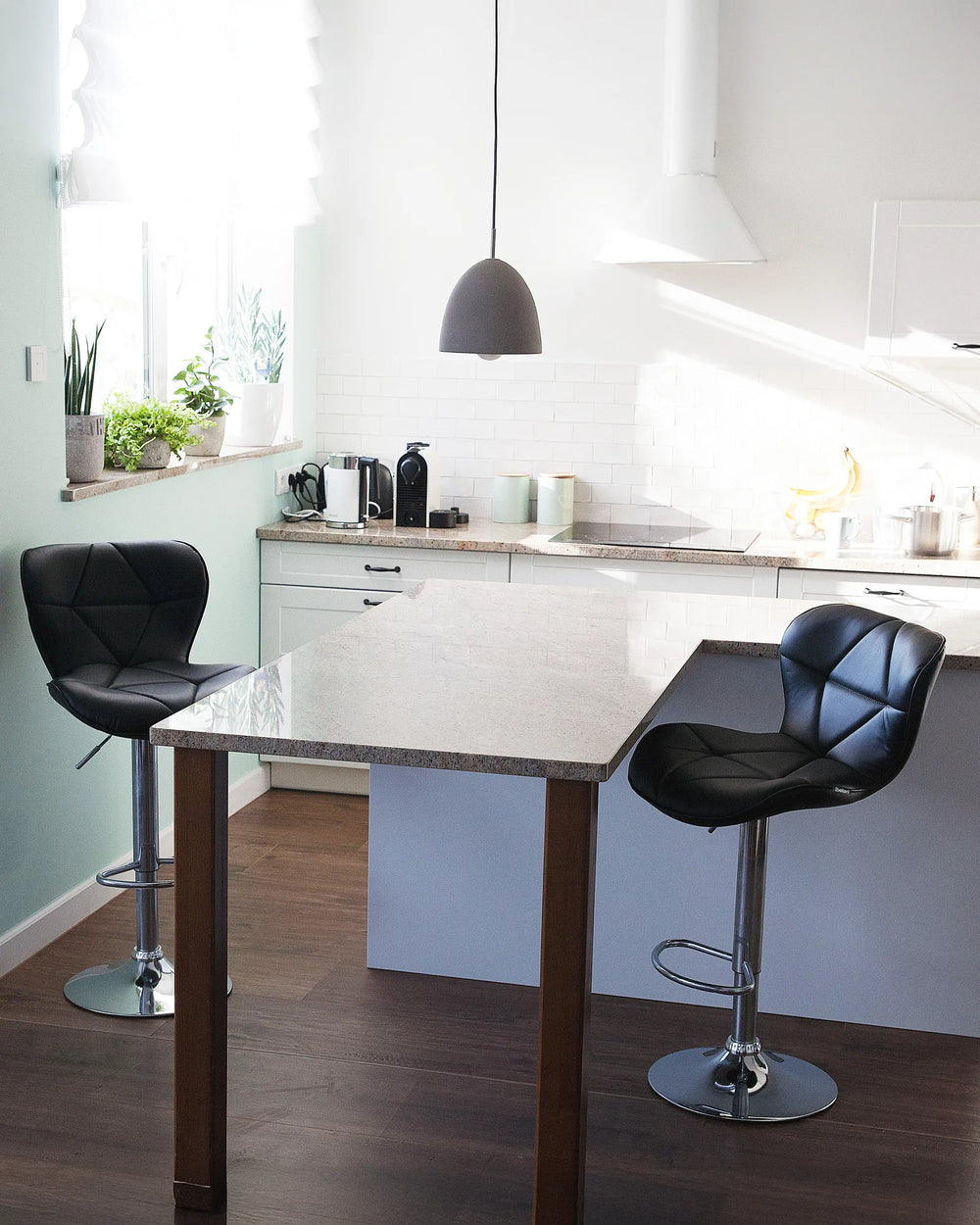 A modern kitchen island with a light stone countertop and dark wood legs, featuring two black tufted bar stools in a bright kitchen setting.