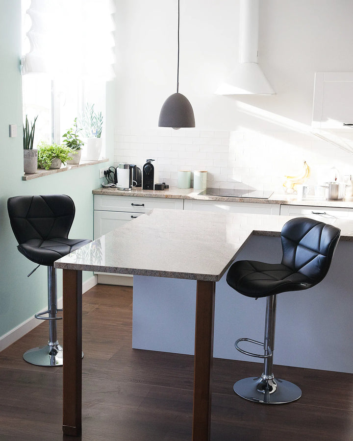 A modern kitchen island with a light stone countertop and dark wood legs, featuring two black tufted bar stools in a bright kitchen setting.