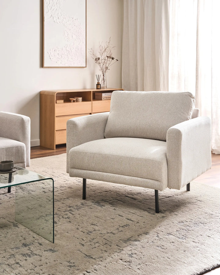 A modern light beige upholstered armchair with black metal legs, positioned in a bright, contemporary living room setting with a glass coffee table, patterned rug, and wooden cabinet in the background.