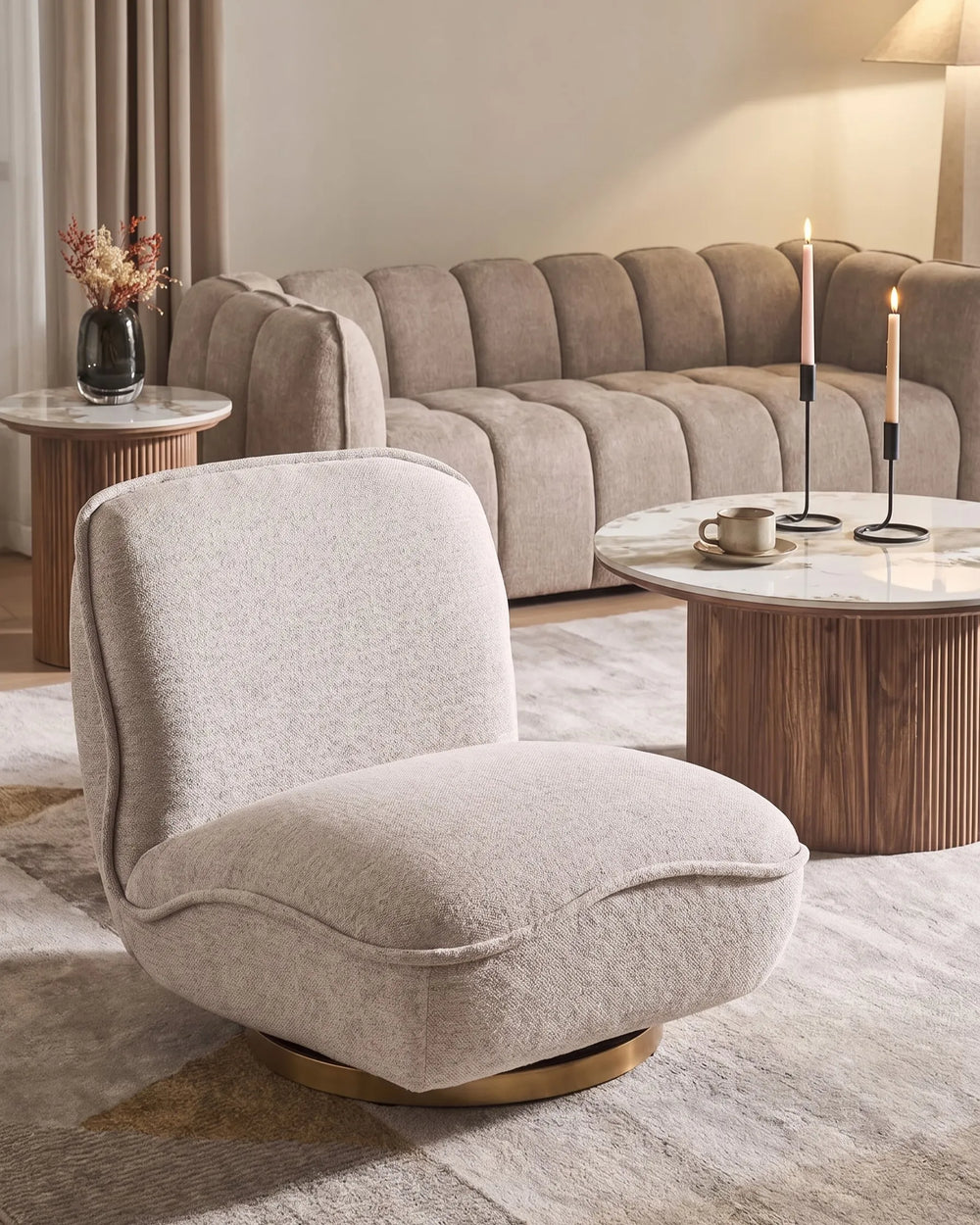 A plush, modern accent chair in a light beige textured fabric with a gold circular swivel base, styled in a warm, contemporary living room setting.