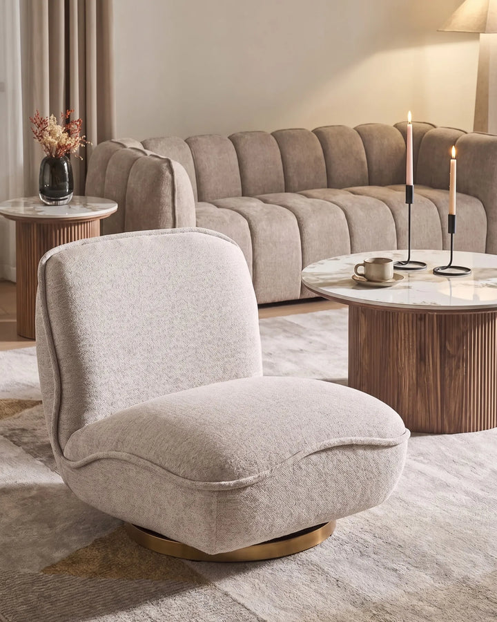 A plush, modern accent chair in a light beige textured fabric with a gold circular swivel base, styled in a warm, contemporary living room setting.