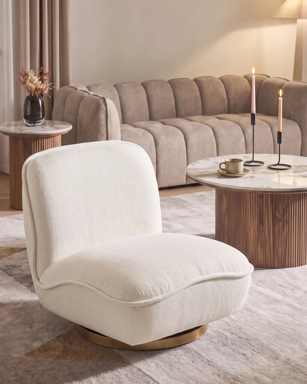 A modern cream-colored upholstered accent chair with a curved back and a round wooden base, set in a stylishly decorated living room with a tufted sofa and marble coffee tables.