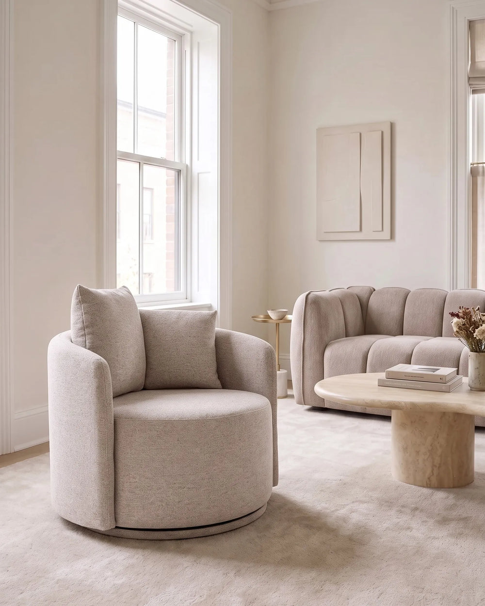 A modern living room featuring a round upholstered swivel armchair in a light beige fabric in the foreground, with a matching beige sofa, a round wooden coffee table, and a large window in the background.