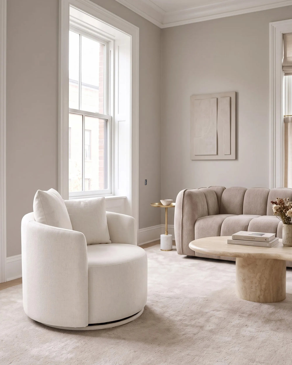 A modern living room featuring a round off-white upholstered swivel armchair in the foreground, with a beige tufted sofa, round wooden coffee table, and light carpet against neutral beige walls.