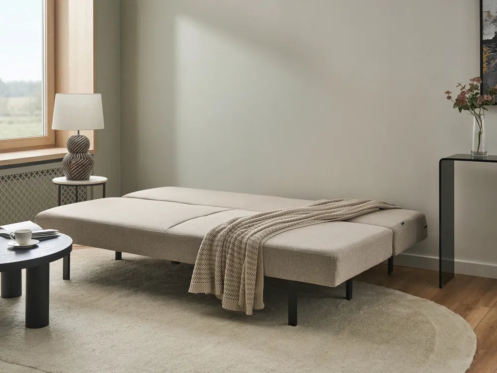 A light beige upholstered daybed or sofa bed with black metal legs, shown folded flat in a modern, neutral-toned living room setting with a textured throw blanket draped over the front.