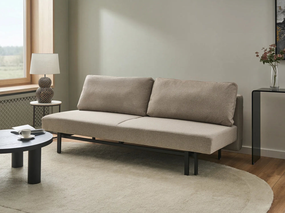A modern two-seat sofa in a warm taupe-beige fabric with a black metal frame, shown in a styled living room setting with a round coffee table, side table, lamp, and rug.