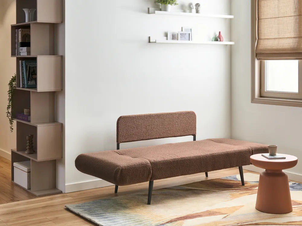 A brown textured fabric daybed with slim black metal legs, styled in a modern living room setting with a matching rug, a small terracotta side table, and a tall shelving unit against a white wall.