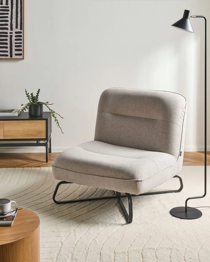 A light beige upholstered accent chair with black metal legs sits on a round woven jute rug in a bright room.