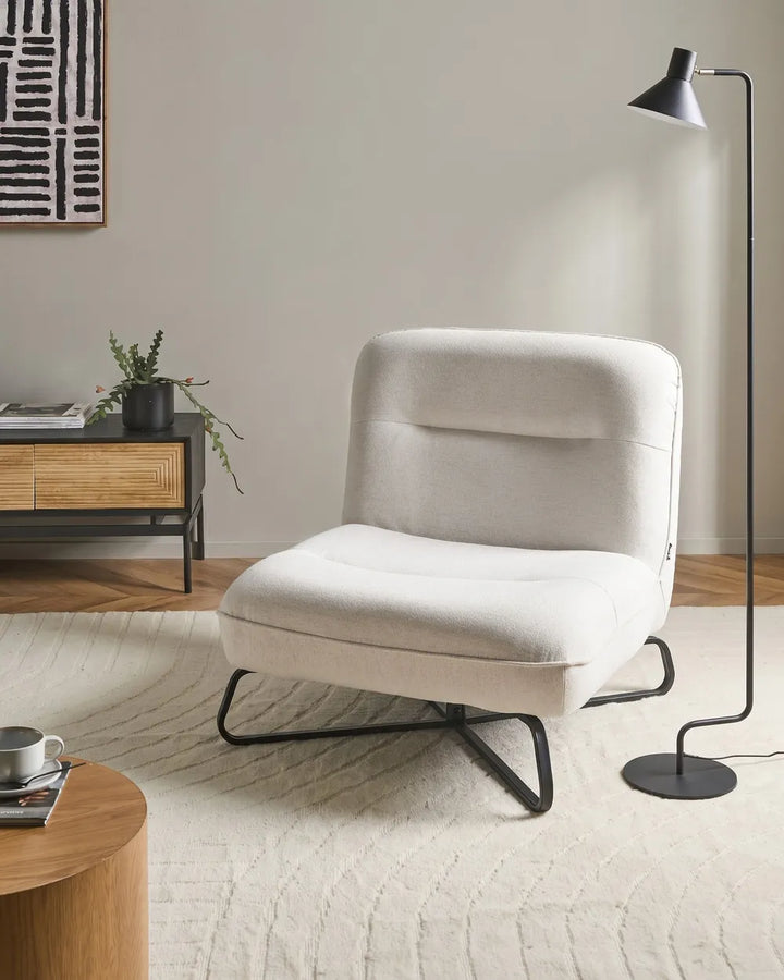A modern upholstered lounge chair in a warm off-white fabric with black metal legs, styled in a minimalist living room next to a black floor lamp.