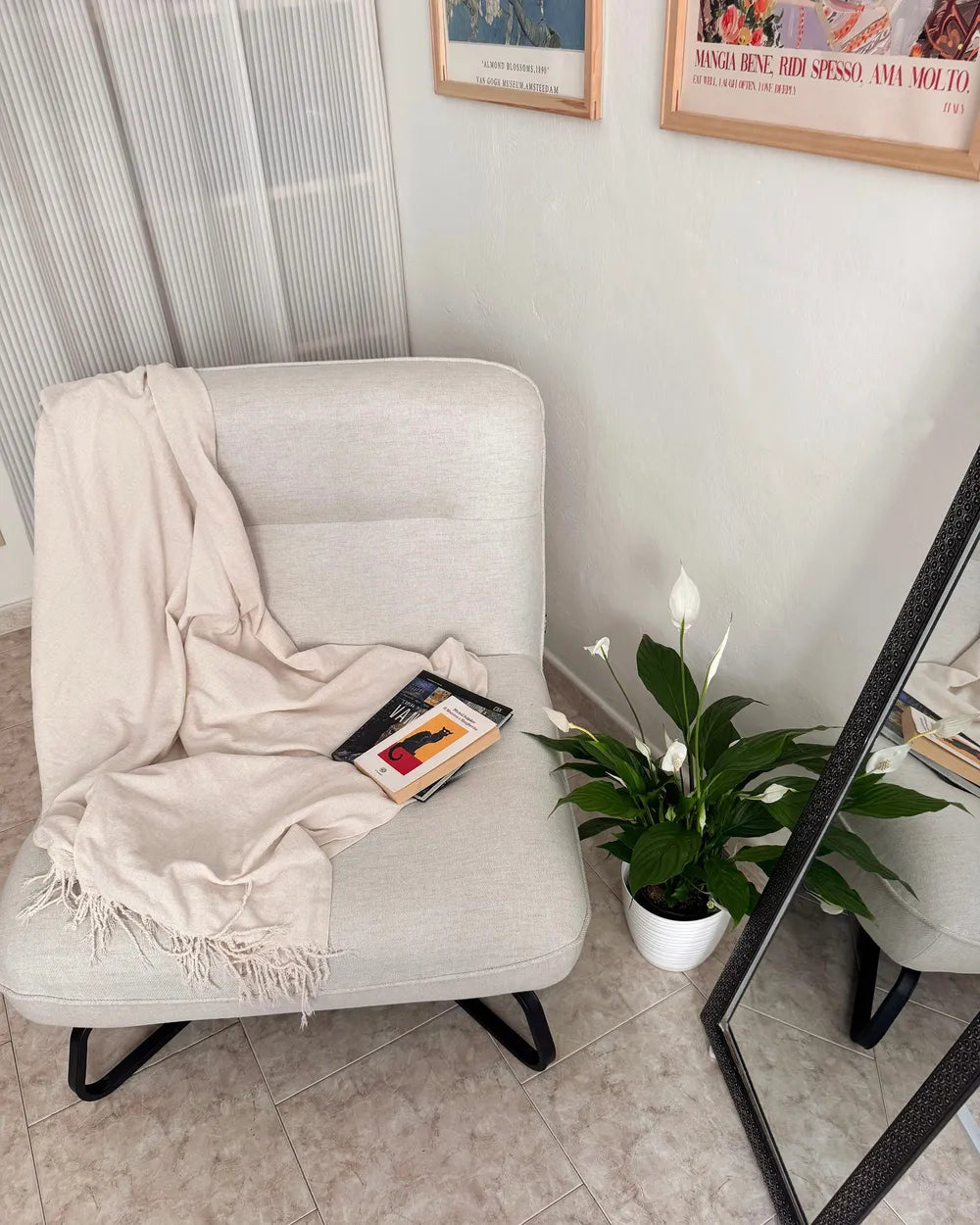 A light beige upholstered armchair in a styled room corner, draped with a cream throw blanket and a book, next to a potted white calla lily plant and a floor mirror.