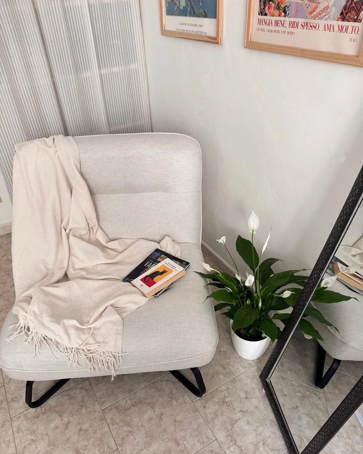 A light beige upholstered armchair in a styled room corner, draped with a cream throw blanket and a book, next to a potted white calla lily plant and a floor mirror.