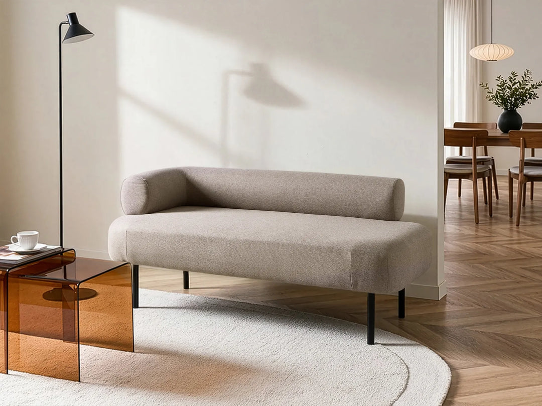 A modern beige upholstered daybed with black metal legs, styled in a minimalist living room setting with a glass side table, floor lamp, and wooden flooring.