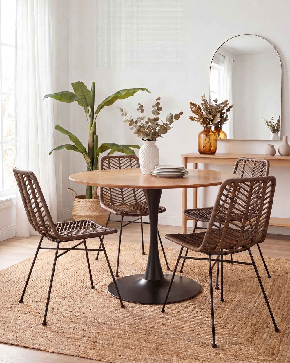 A round wooden dining table with a black pedestal base, surrounded by four woven brown chairs with black metal legs, set on a jute rug in a bright, modern dining room.