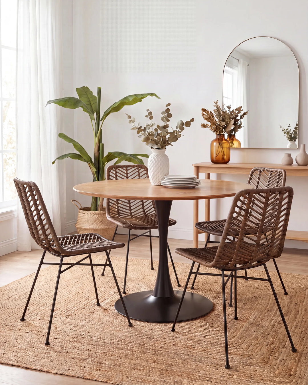 A round wooden dining table with a black pedestal base, surrounded by four woven brown chairs with black metal legs, set on a jute rug in a bright, modern dining room.