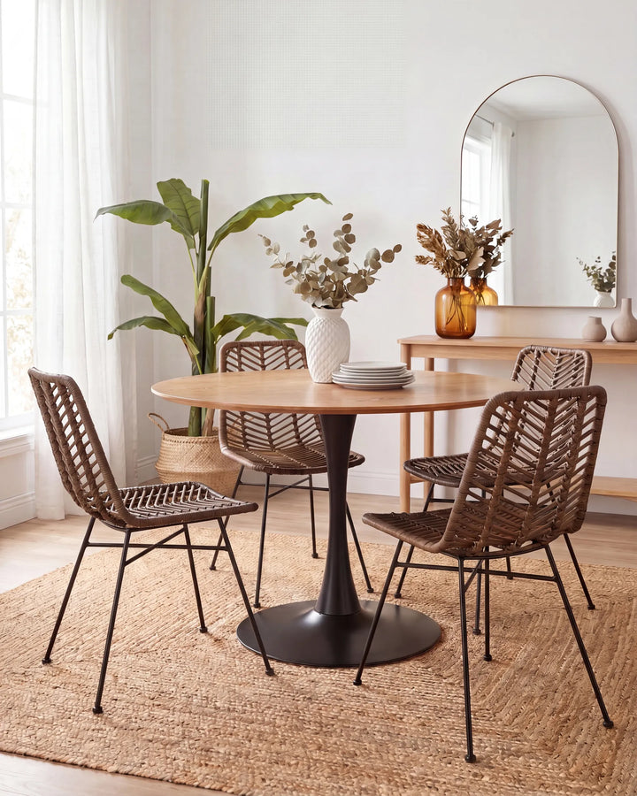 A round wooden dining table with a black pedestal base, surrounded by four woven brown chairs with black metal legs, set on a jute rug in a bright, modern dining room.