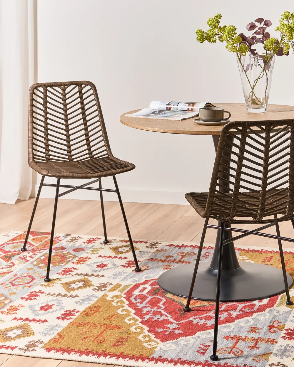 Two wicker chairs at a round wooden table with a colorful rug underneath.