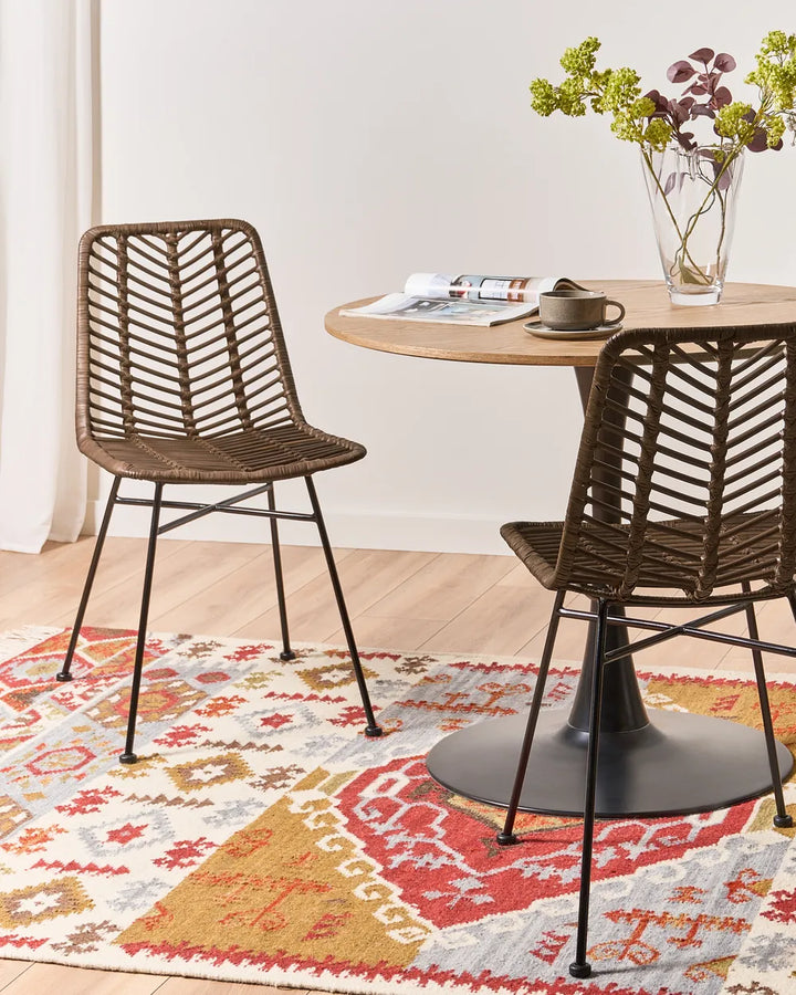 Two wicker chairs at a round wooden table with a colorful rug underneath.