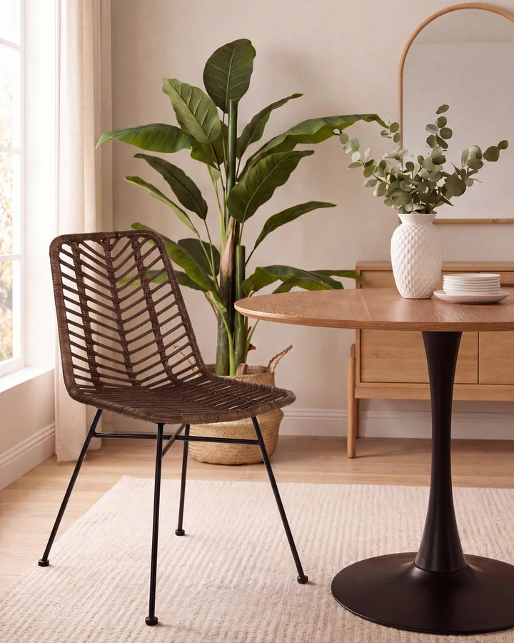 A brown woven rattan dining chair with black metal legs, positioned next to a round wooden table in a bright, modern dining room with a potted plant and a white vase in the background.