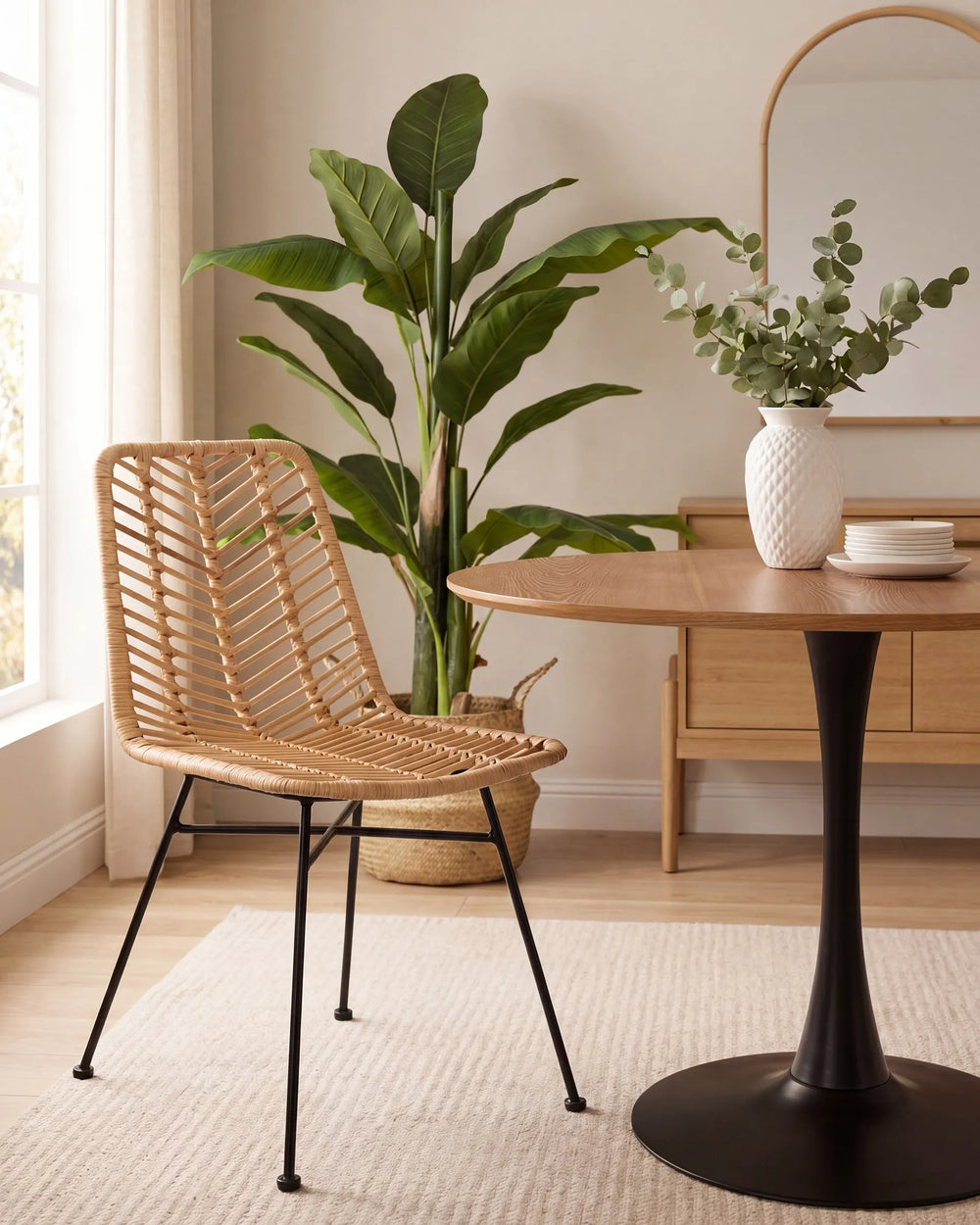 A woven rattan dining chair with black metal legs sits beside a round wooden table in a bright, modern room with a potted plant and a white vase.