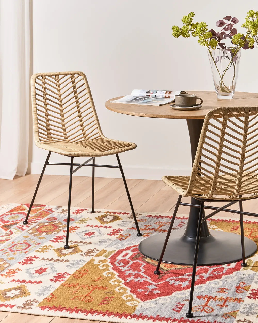 Two woven rattan dining chairs with black metal legs arranged around a round wooden table on a patterned area rug in a bright room.