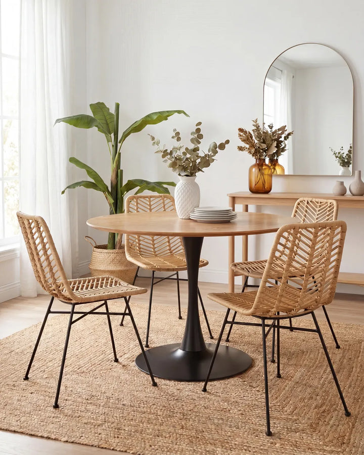 A round wooden dining table with a black pedestal base, surrounded by four woven rattan chairs with black metal legs, set on a jute rug in a bright, modern dining room.