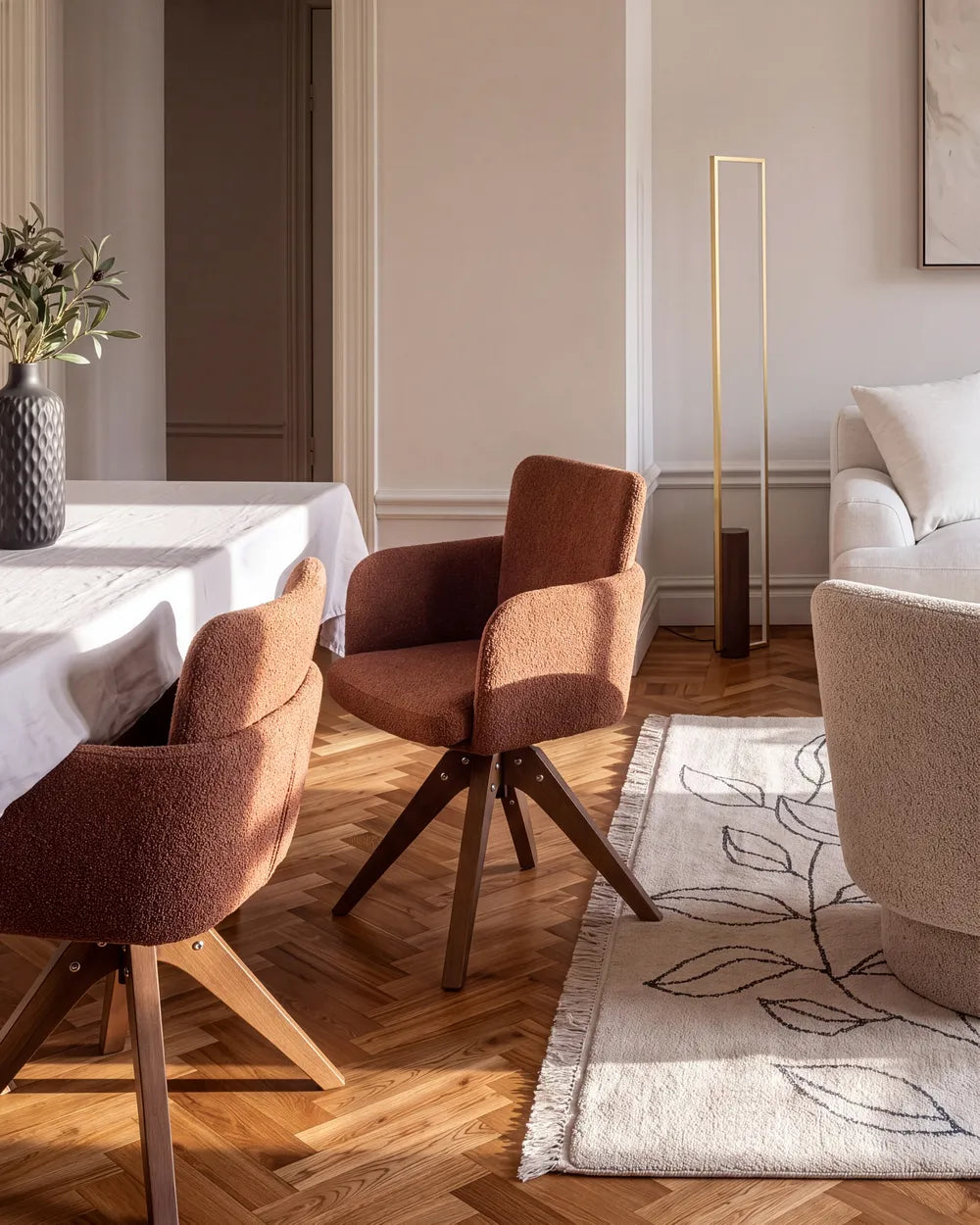 A modern dining room scene featuring two upholstered swivel chairs in a warm rust-brown fabric with dark wood legs, set on a herringbone wood floor next to a white table and a patterned rug.