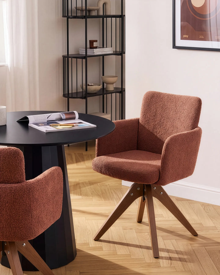 A modern upholstered dining chair in a warm rust-brown bouclé fabric with a wooden swivel base, shown in a styled dining room setting next to a black round table.