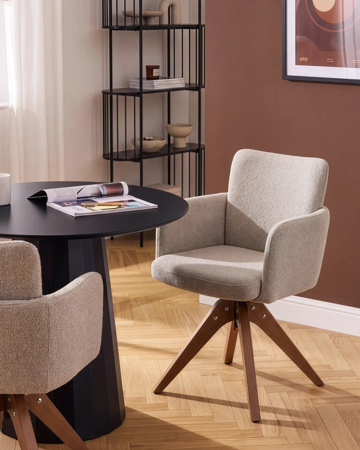 A modern upholstered dining chair with a textured light beige fabric and a four-star wooden swivel base, shown in a styled dining room setting next to a black round table.