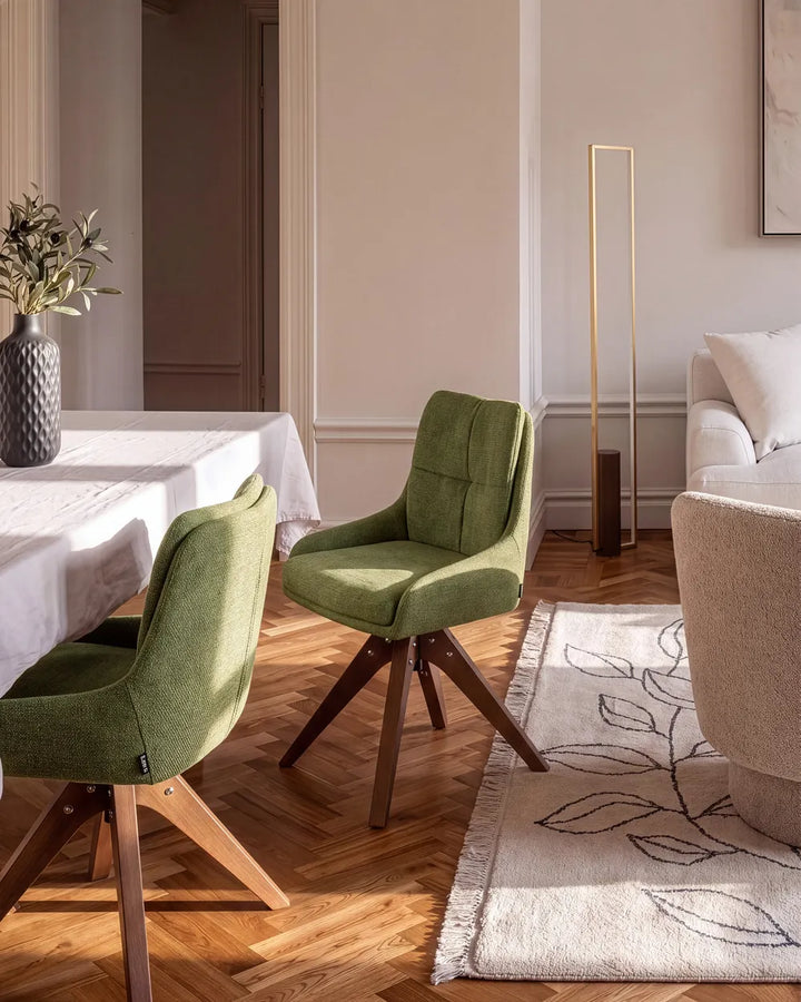 A modern dining room scene featuring two green upholstered swivel chairs with wooden legs, set on a herringbone wood floor next to a dining table and a cream-colored sofa.