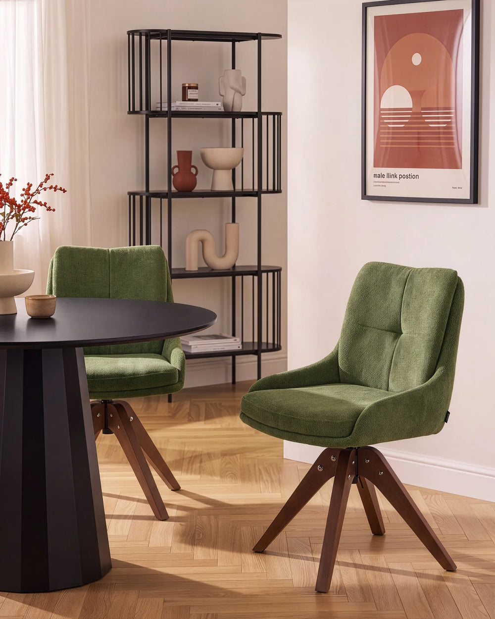 A modern dining room scene featuring two green velvet swivel chairs with dark wood star legs, set around a black round table. A tall black metal shelving unit stands against the wall, and a framed art print hangs on the wall.