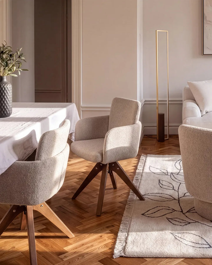 A modern dining room scene featuring two upholstered swivel chairs with textured light beige fabric and dark wood legs, set on a herringbone wood floor next to a dining table and a patterned rug.