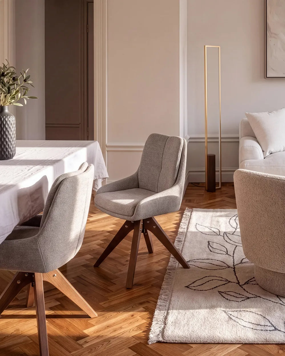 A modern dining chair with light gray upholstered fabric and dark wood legs, shown in a styled dining room setting with a table, sofa, rug, and warm wood flooring.