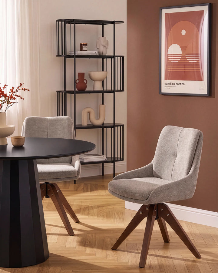 A modern dining room scene featuring a light gray upholstered swivel chair with dark wooden legs, a matching gray chair, and a black round table. A black metal shelving unit stands against a warm brown wall with framed wall art and decorative objects.