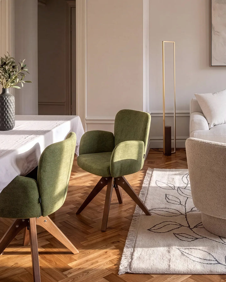 Two green upholstered swivel dining chairs with wooden legs in a bright, modern dining room with herringbone wood floors and a white table runner.
