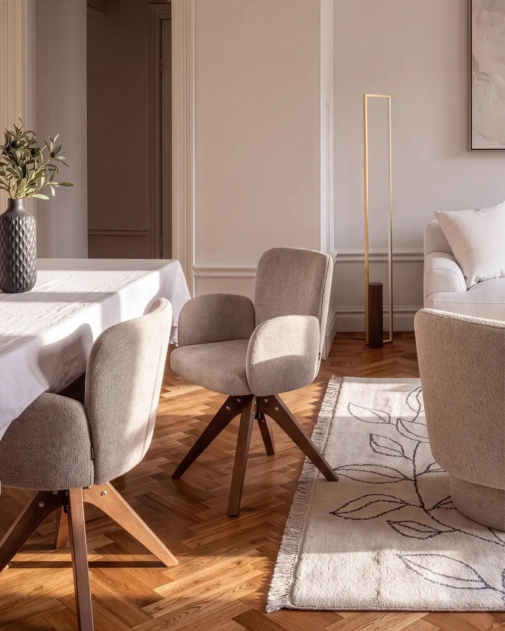 A modern dining room scene featuring upholstered swivel dining chairs with light beige fabric and dark wood legs, arranged around a white table on a herringbone wood floor. A cream rug with a leaf pattern, a floor lamp, and a sofa are visible in the background.