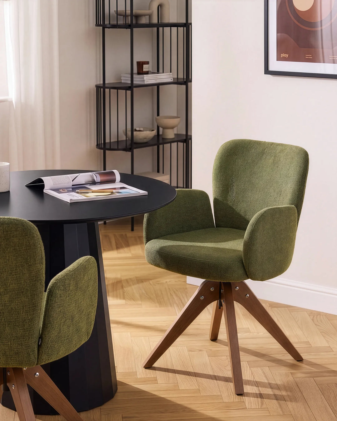 An olive green upholstered swivel armchair with wooden legs, shown in a modern dining room setting next to a black round table.