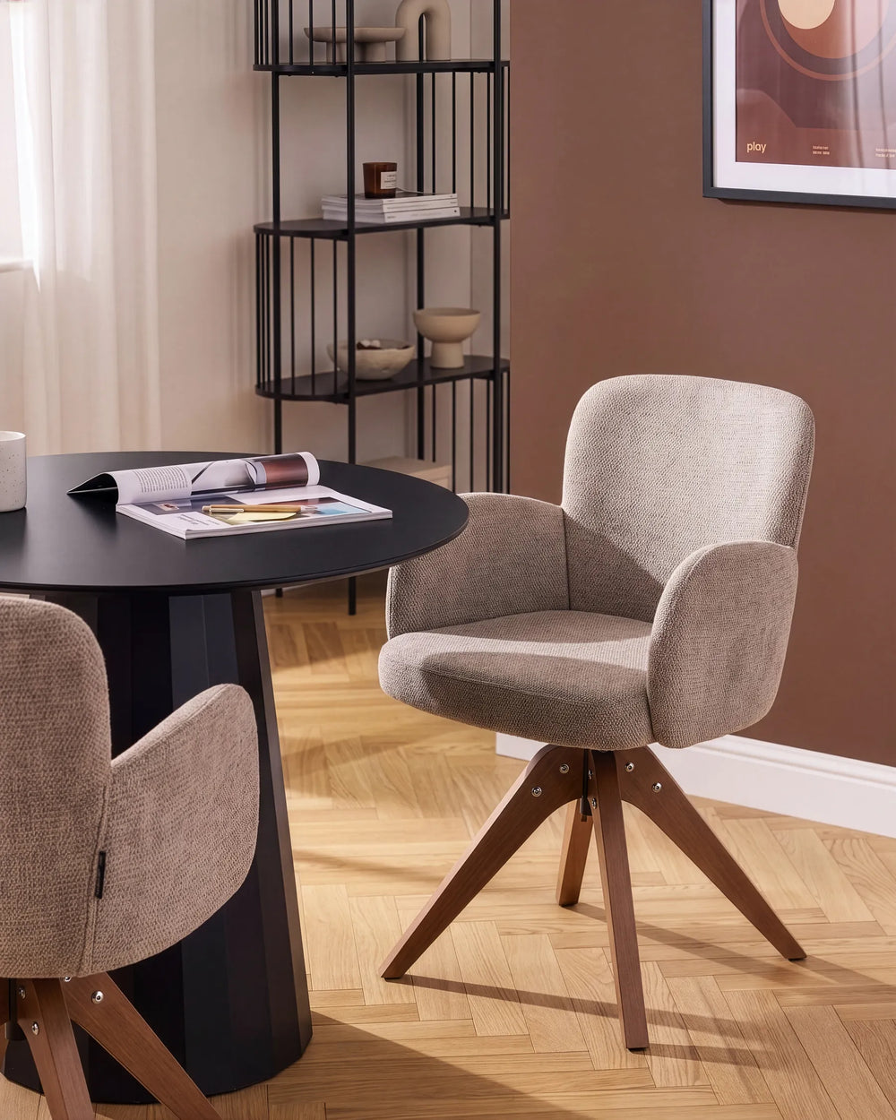 A modern upholstered dining chair in a warm taupe fabric with a wooden swivel base, shown in a styled dining room setting next to a black round table.