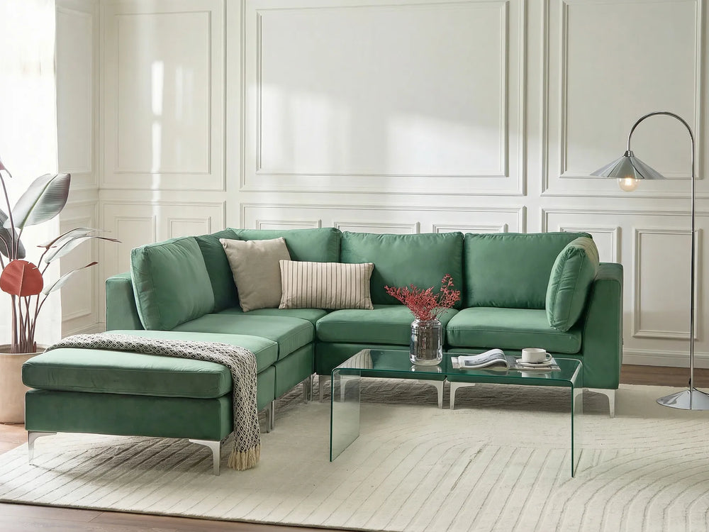 A modern green sectional sofa with an ottoman, styled in a bright living room with a glass coffee table, neutral throw pillows, a patterned rug, and a floor lamp.