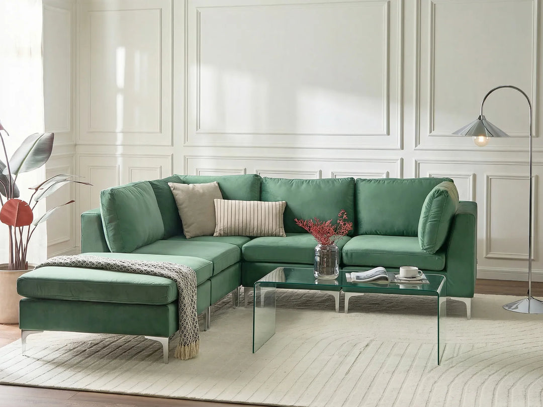 A modern green sectional sofa with an ottoman, styled in a bright living room with a glass coffee table, neutral throw pillows, a patterned rug, and a floor lamp.