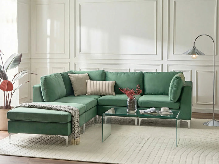 A modern green sectional sofa with an ottoman, styled in a bright living room with a glass coffee table, neutral throw pillows, a patterned rug, and a floor lamp.