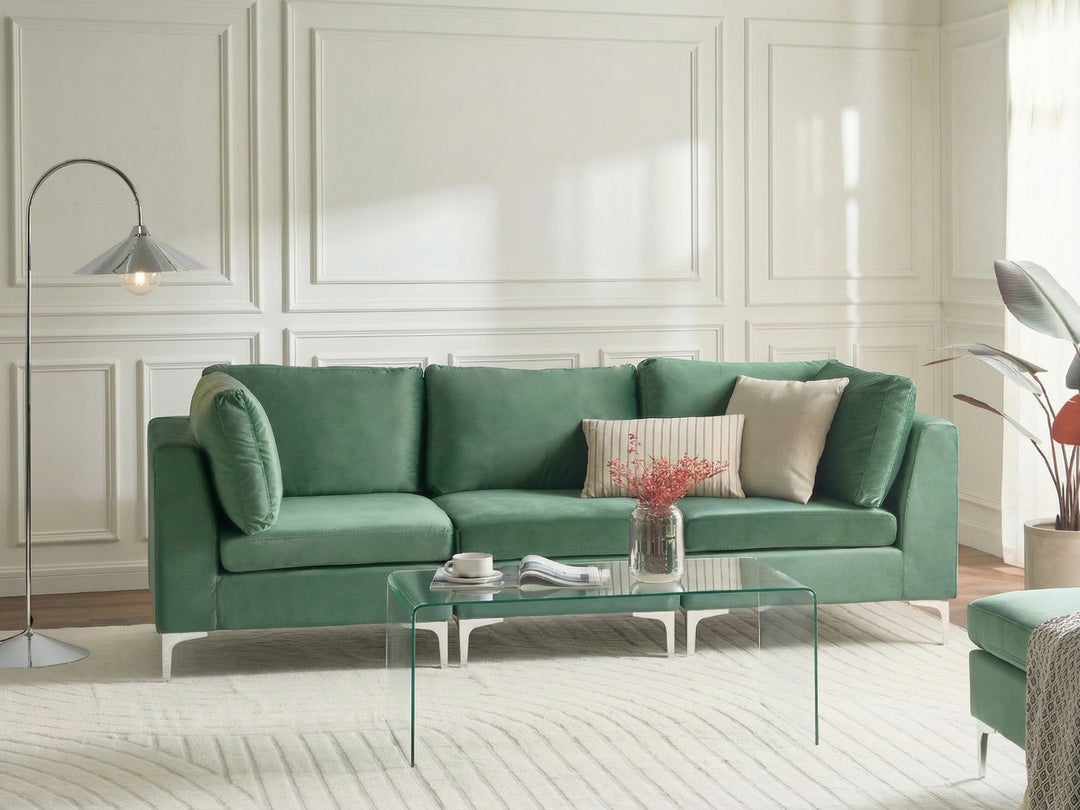 A green upholstered three-seat sofa in a bright, modern living room with white paneled walls, a glass coffee table, and a floor lamp.