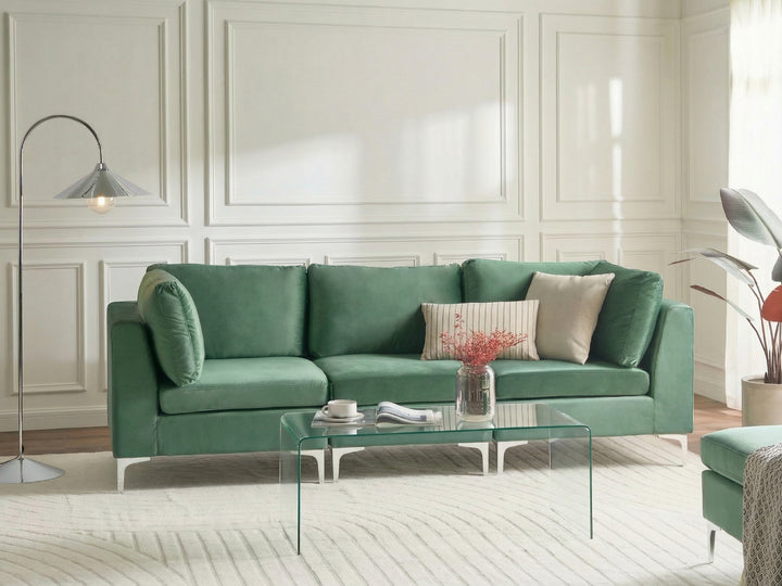 A green upholstered three-seat sofa in a bright, modern living room with white paneled walls, a glass coffee table, and a floor lamp.