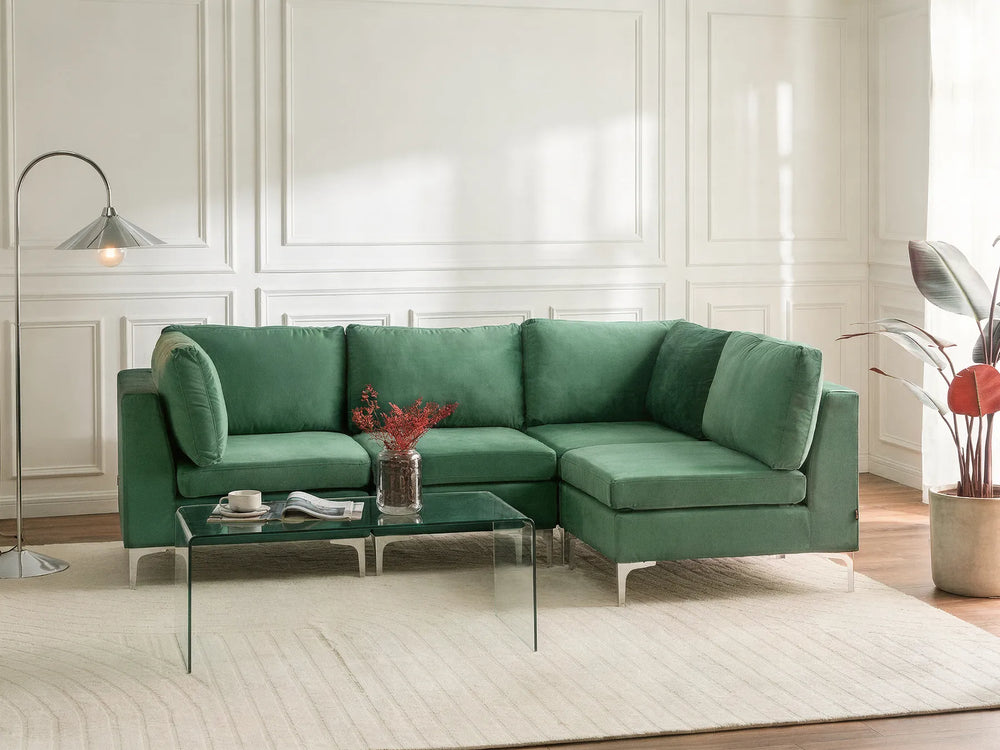 A modern green sectional sofa with a left-facing chaise lounge, styled in a bright living room with a glass coffee table, a floor lamp, and a potted plant.