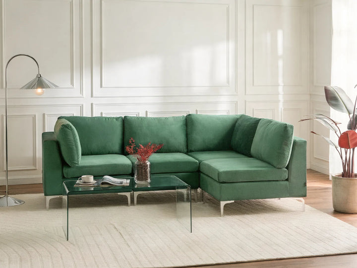 A modern green sectional sofa with a left-facing chaise lounge, styled in a bright living room with a glass coffee table, a floor lamp, and a potted plant.
