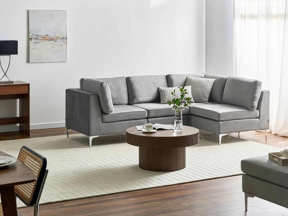 A modern gray fabric sectional sofa with a chaise lounge, styled in a bright, contemporary living room with a round wooden coffee table, textured cream rug, and wood flooring.