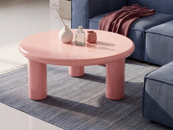 A glossy pink round coffee table with thick cylindrical legs, styled in a modern living room setting on a blue-gray rug next to a blue sofa.