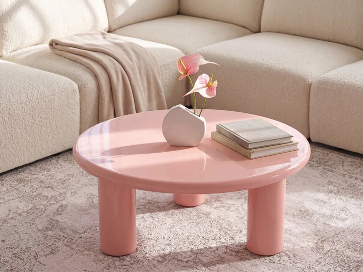 A round, low-profile coffee table in glossy pastel pink, styled in a living room setting with a beige boucle sofa, a draped throw blanket, a small white vase with pink flowers, and a stack of books on top.