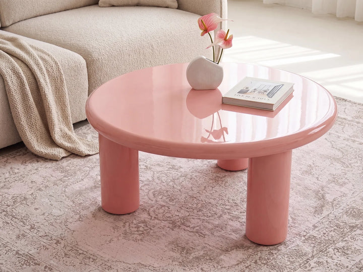 A round low coffee table in glossy pastel pink with chunky cylindrical legs, styled in a bright living room with a beige sofa, patterned rug, vase of flowers, and a book on top.