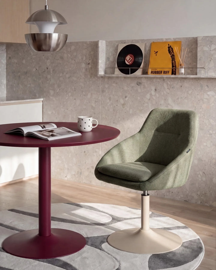 A modern swivel chair in sage green upholstery with a cream pedestal base, styled in a contemporary room next to a round burgundy table.