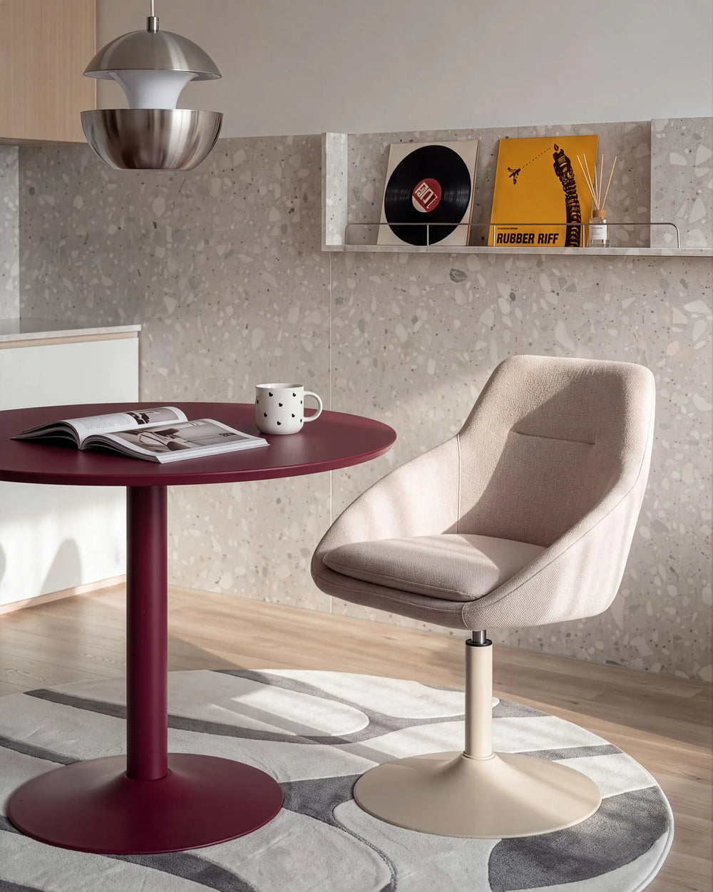 A modern interior scene featuring a beige upholstered swivel chair beside a round dark red table, with a textured stone wall, shelf decor, and a hanging pendant light in the background.